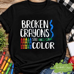 May include: A black t-shirt featuring the white text BROKEN CRAYONS STILL COLOR. The design includes a row of five vertical crayons in red, orange, yellow, green, and blue, a single horizontal green broken crayon, and a blue wavy line graphic.