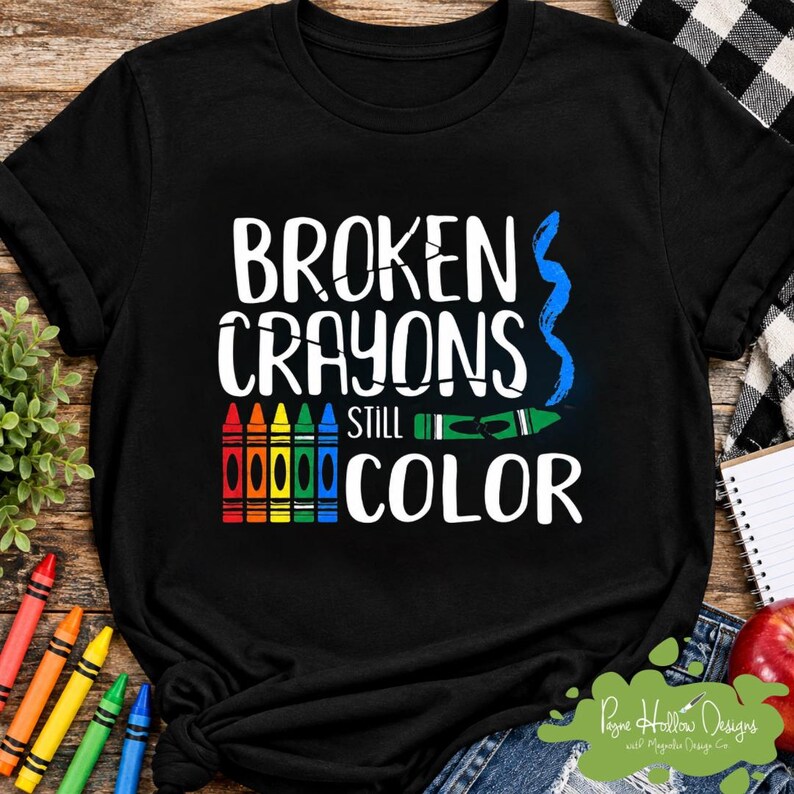 May include: A black t-shirt featuring the white text BROKEN CRAYONS STILL COLOR. The design includes a row of five vertical crayons in red, orange, yellow, green, and blue, a single horizontal green broken crayon, and a blue wavy line graphic.