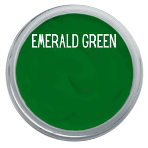 May include: A circular container filled with vibrant emerald green paint. The text EMERALD GREEN is printed in white capital letters across the top center of the smooth, saturated green surface, which is enclosed by a clear plastic rim.