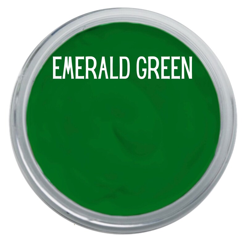 May include: A circular container filled with vibrant emerald green paint. The text EMERALD GREEN is printed in white capital letters across the top center of the smooth, saturated green surface, which is enclosed by a clear plastic rim.