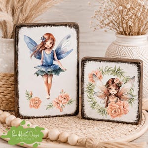 May include: Two rectangular wooden signs with distressed dark edges. The taller sign features a red-haired fairy with blue wings and a blue dress. The shorter sign displays a fairy with light wings surrounded by a wreath of peach-colored flowers.