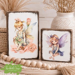 May include: Two rectangular wooden blocks with distressed dark edges featuring watercolor illustrations of fairies. The taller block shows a fairy with a pink rose, while the shorter block displays a fairy with purple wings. Both sit on a wooden bead garland.