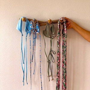 May include: A selection of swimwear tops and accessories suspended from a wooden rack. The items include a light blue top, a blue floral print top, a green crochet top with seashells, and two floral print straps. They are displayed against a white wall.