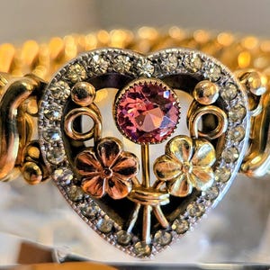 May include: A gold-toned stretch bracelet featuring a central heart-shaped charm. The charm is bordered by small clear crystals and contains a single round pink stone above two small metal flowers, one rose gold-toned and one yellow gold-toned.