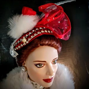 May include: A porcelain doll with red hair wearing a white fur coat and a red and white hat with rhinestones and a white feather.