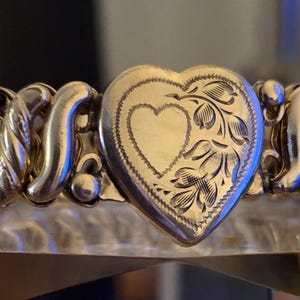 May include: A gold-toned bracelet with a heart-shaped centerpiece. The heart is engraved with floral designs and a smaller heart in the center. The bracelet has an expandable design, with a series of linked segments.