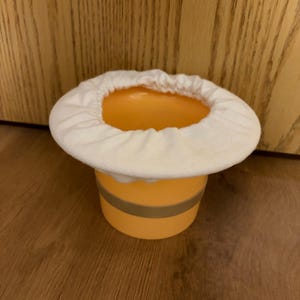 May include: A cylindrical orange plastic container featuring a horizontal gray band around its center. The top opening is covered by a white, elasticized fabric cover that creates a gathered, ruffled edge around the rim of the container.