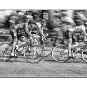 Puede incluir: Tres ciclistas compiten en una carretera pavimentada. Los ciclistas llevan ropa deportiva y cascos. Los ciclistas están borrosos, lo que sugiere velocidad.