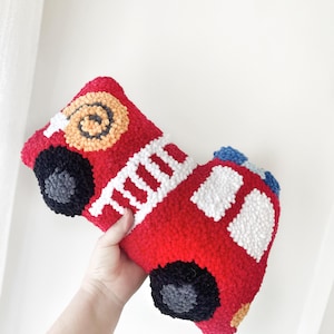 Puede incluir: Una almohada de felpa roja en forma de cami&oacute;n de bomberos con detalles blancos, negros, azules y amarillos. La almohada est&aacute; hecha de hilo y tiene una apariencia texturizada y esponjosa.