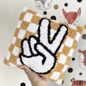 May include: A square decorative pillow with a checkerboard pattern in white and mustard yellow. The pillow features a large, black and white peace sign in the center. The pillow is made of a soft, textured material.