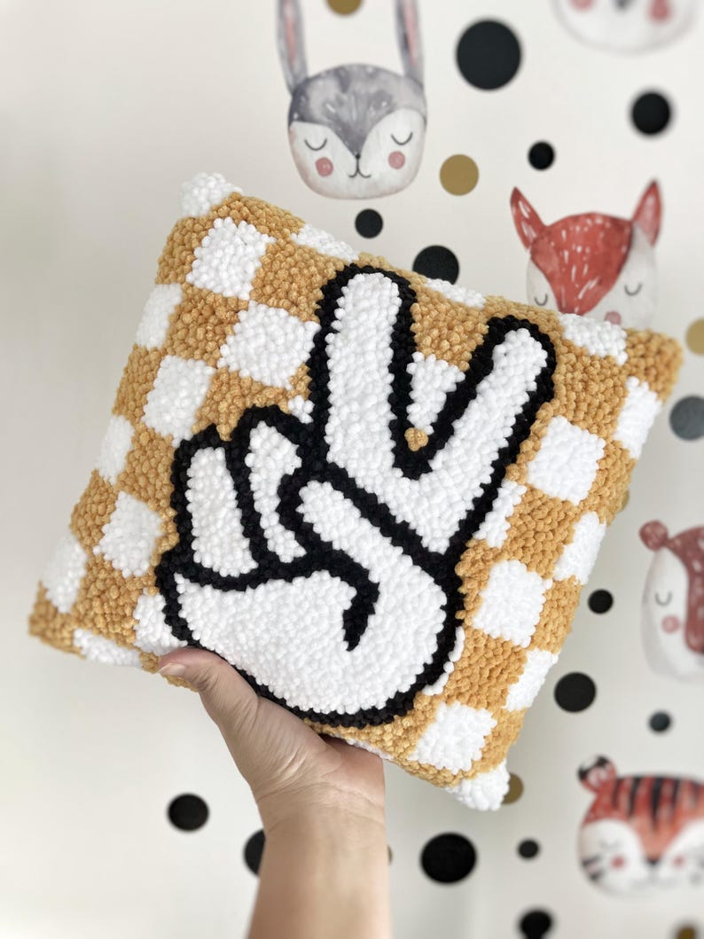 May include: A square decorative pillow with a checkerboard pattern in white and mustard yellow. The pillow features a large, black and white peace sign in the center. The pillow is made of a soft, textured material.