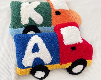 May include: Three colorful, handmade truck-shaped pillows. One is green with a white "K", another is orange, and the third is red and blue with a white "A". Each has black wheels and a white cab.