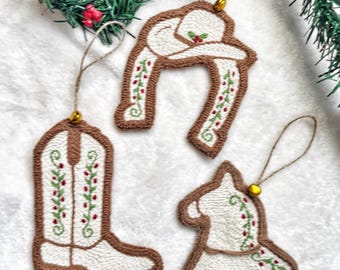 May include: Three punch needle ornaments featuring a cowboy boot, a cowboy hat, and a horse. Each piece is crafted with white and brown yarn, accented with embroidered green vines and red berries, and includes a gold bead and twine loop for hanging.