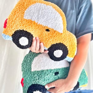 Puede incluir: Dos cojines hechos a mano con forma de coche. El coj&iacute;n superior es amarillo con detalles blancos, negros, rojos y azules. El coj&iacute;n inferior es verde con detalles blancos, negros y rojos. Ambos cojines tienen un aspecto texturizado y afelpado.