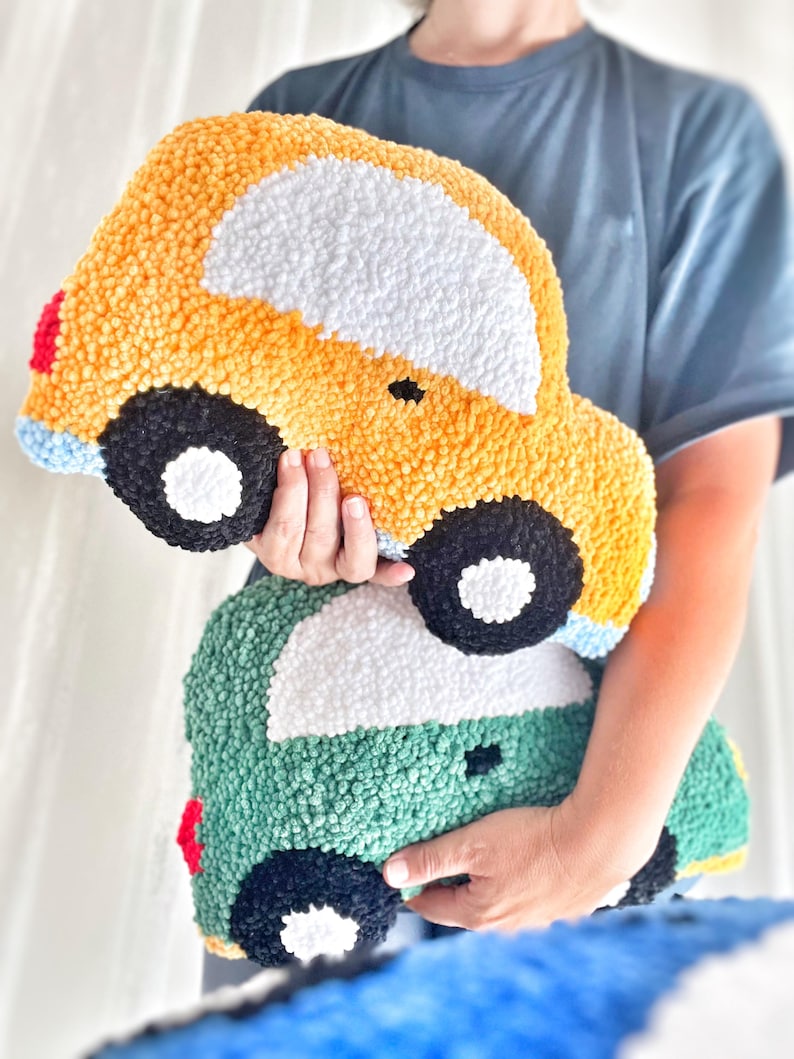 Puede incluir: Dos cojines hechos a mano con forma de coche. El coj&iacute;n superior es amarillo con detalles blancos, negros, rojos y azules. El coj&iacute;n inferior es verde con detalles blancos, negros y rojos. Ambos cojines tienen un aspecto texturizado y afelpado.