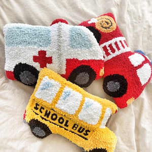 Könnte beinhalten: Drei Plüschtiere in Form von Fahrzeugen: ein gelber Schulbus mit der Aufschrift "SCHOOL BUS" an der Seite, ein rotes Feuerwehrauto mit weißen Streifen und ein weißer Krankenwagen mit einem roten Kreuz.