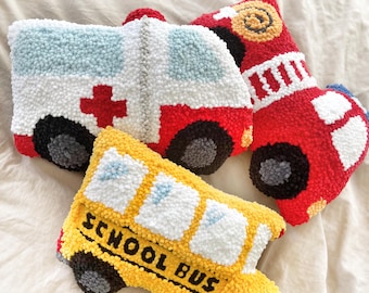 May include: Three plush toys shaped like vehicles: a yellow school bus with the words "SCHOOL BUS" on the side, a red fire truck with white stripes, and a white ambulance with a red cross.