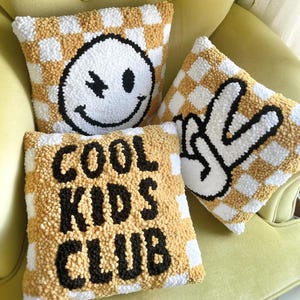 May include: Three decorative throw pillows with a checkerboard pattern in white and tan. One pillow features a winking smiley face, another a peace sign, and the third reads "COOL KIDS CLUB" in black block letters. The pillows are on a yellow chair.