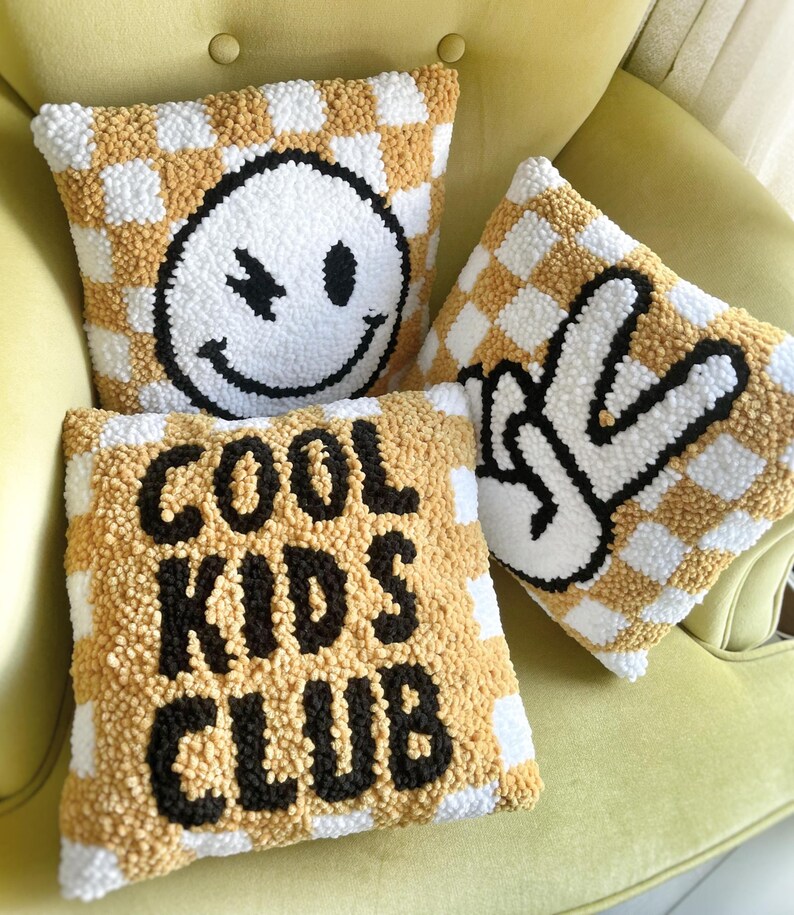 May include: Three decorative throw pillows with a checkerboard pattern in white and tan. One pillow features a winking smiley face, another a peace sign, and the third reads "COOL KIDS CLUB" in black block letters. The pillows are on a yellow chair.