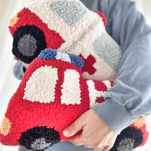 Puede incluir: Un juguete de peluche rojo y blanco con forma de cami&oacute;n de bomberos. El juguete est&aacute; hecho de hilo y tiene un efecto 3D. El juguete est&aacute; siendo sostenido por la mano de una persona.