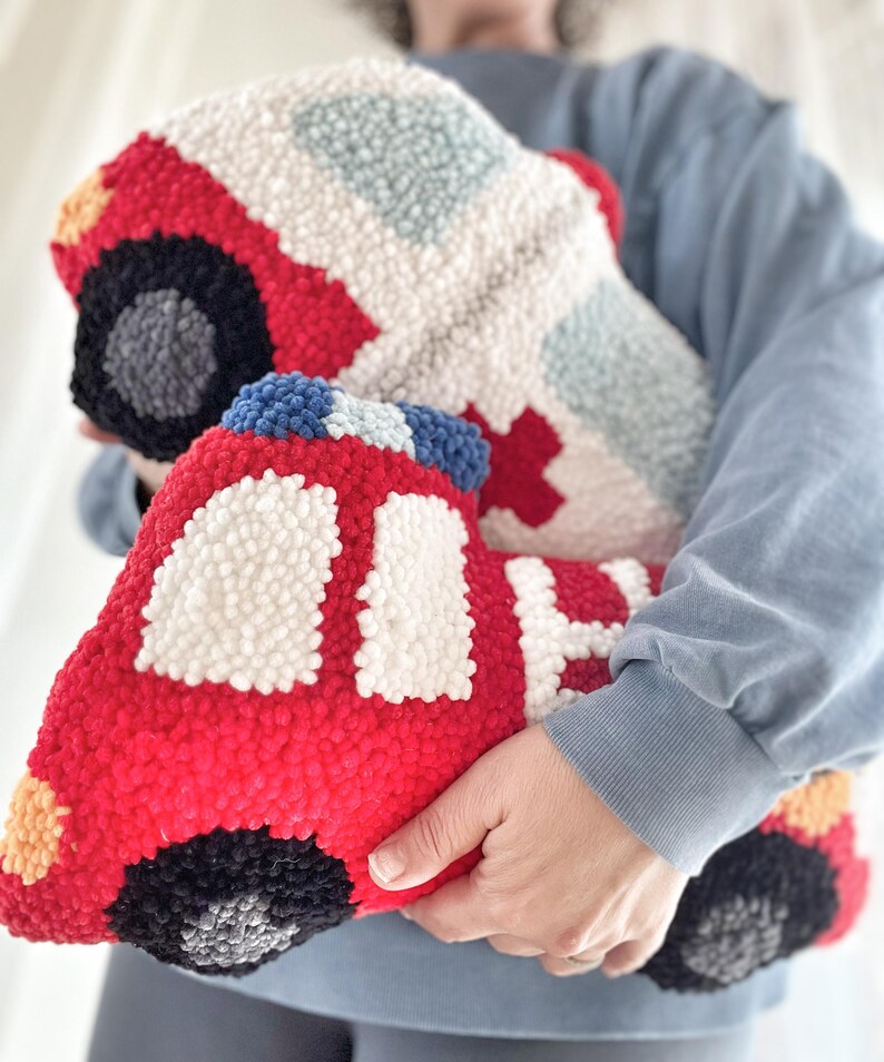 Puede incluir: Un juguete de peluche rojo y blanco con forma de cami&oacute;n de bomberos. El juguete est&aacute; hecho de hilo y tiene un efecto 3D. El juguete est&aacute; siendo sostenido por la mano de una persona.