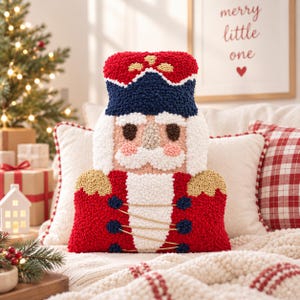 May include: A punch needle throw pillow shaped like a nutcracker soldier. The pillow features a red hat with gold accents, a dark blue brim, a white beard, and a red uniform jacket with gold trim, dark blue buttons, and gold thread details.