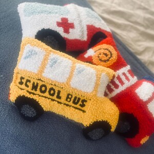 Puede incluir: Tres veh&iacute;culos de peluche: un autob&uacute;s escolar amarillo con las palabras "SCHOOL BUS" en el lateral, un cami&oacute;n de bomberos rojo con una cruz blanca en el lateral y una ambulancia blanca con una cruz roja en el lateral.