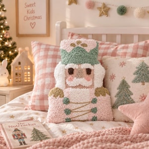 May include: A punch needle decorative pillow shaped like a nutcracker, featuring a soft pastel color palette of pink, sage green, and white. The design includes a detailed uniform with gold thread accents, a crown, and a textured, tufted surface.