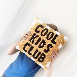 May include: A square, textured pillow with the words "COOL KIDS CLUB" in black letters on a yellow background. The pillow has a white border with yellow stripes. The pillow is being held by a person wearing a blue shirt.