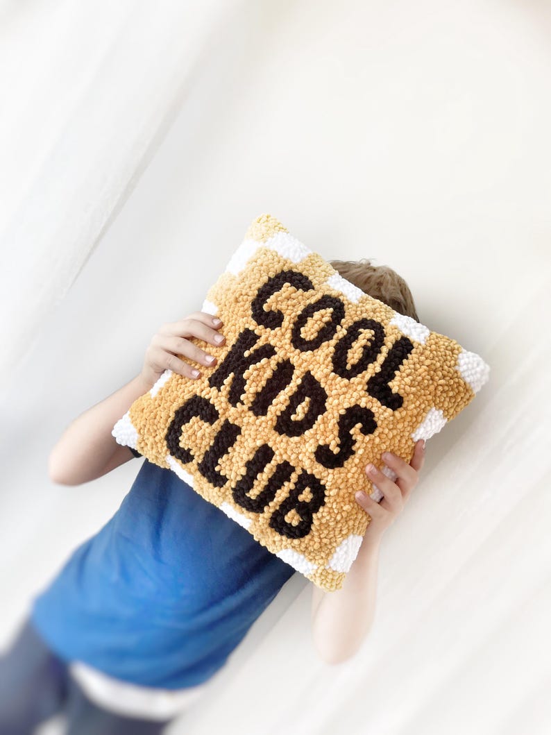 May include: A square, textured pillow with the words "COOL KIDS CLUB" in black letters on a yellow background. The pillow has a white border with yellow stripes. The pillow is being held by a person wearing a blue shirt.