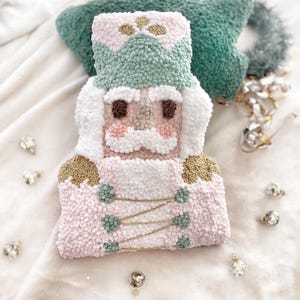 May include: A punch needle nutcracker decoration featuring a soft pastel color palette of light pink, sage green, white, and gold accents. The figure has a textured, looped yarn surface, a tall hat, a mustache, and a uniform with gold thread details.