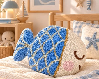 May include: A plush fish-shaped pillow featuring a textured blue and white pom-pom scale pattern, a cream-colored face with closed brown eyes, and a soft pink cheek. The pillow has a distinct tail fin and a rounded body design made of yarn.