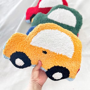 Puede incluir: Una colecci&oacute;n de cojines con forma de coche en varios colores. Los cojines son amarillos, verdes y rojos, con ventanas blancas y ruedas negras y blancas. El material de felpa les da un aspecto texturizado.