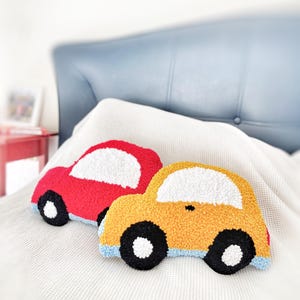Puede incluir: Dos cojines con forma de coche, uno rojo y otro amarillo. Ambos tienen detalles de ventanas blancas, ruedas negras y ribetes azules. Los cojines est&aacute;n sobre una manta blanca, con un cabecero azul al fondo.