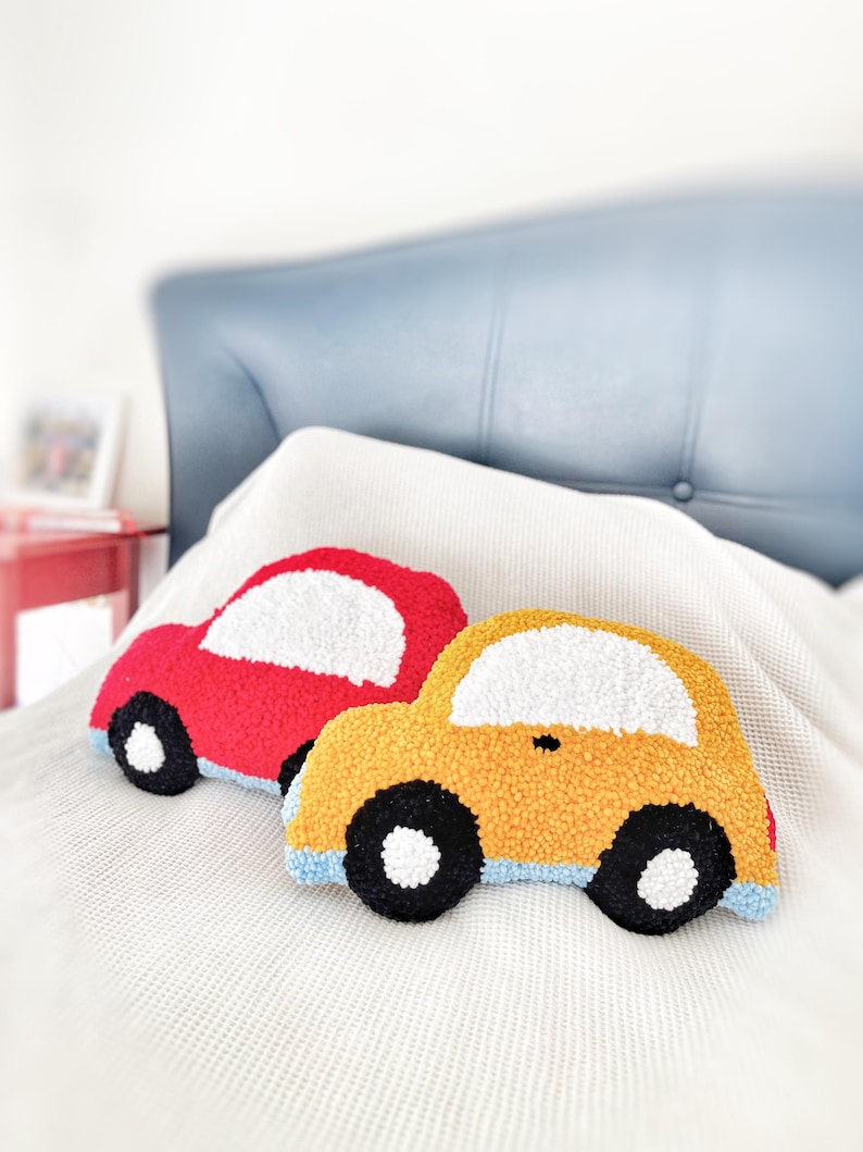 Puede incluir: Dos cojines con forma de coche, uno rojo y otro amarillo. Ambos tienen detalles de ventanas blancas, ruedas negras y ribetes azules. Los cojines est&aacute;n sobre una manta blanca, con un cabecero azul al fondo.