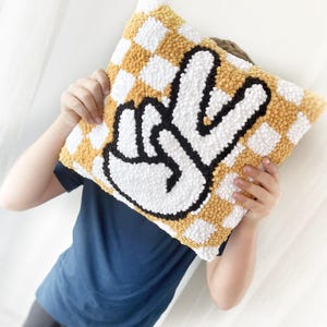 May include: Square decorative pillow with a checkerboard pattern in yellow and white. The pillow features a large peace sign in white with a black outline. The pillow is being held up by a person.