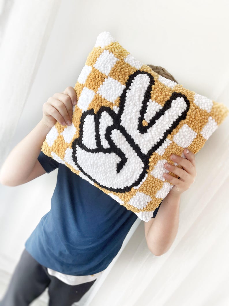 May include: Square decorative pillow with a checkerboard pattern in yellow and white. The pillow features a large peace sign in white with a black outline. The pillow is being held up by a person.