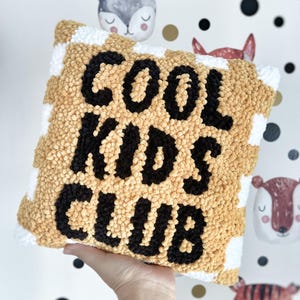 May include: A square, textured pillow with the words "COOL KIDS CLUB" in black lettering. The pillow has a tan and white checkered pattern. The background features a whimsical wallpaper with animal faces.