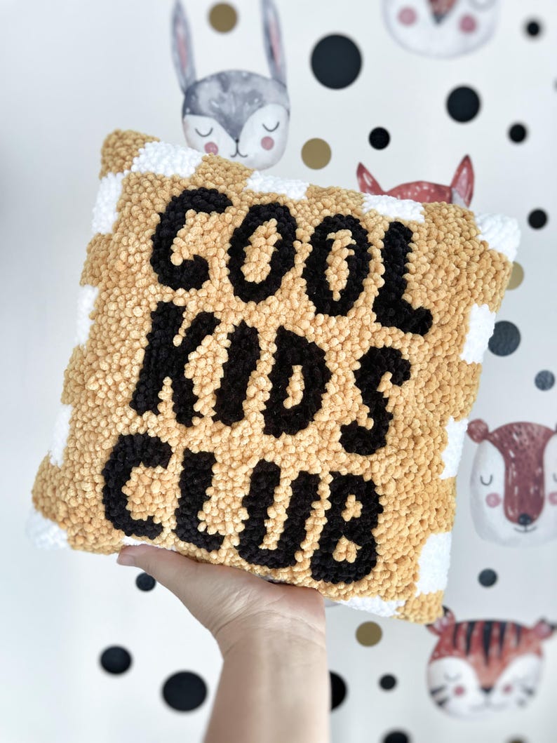 May include: A square, textured pillow with the words "COOL KIDS CLUB" in black lettering. The pillow has a tan and white checkered pattern. The background features a whimsical wallpaper with animal faces.