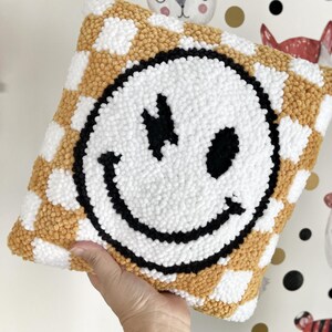 May include: A square, handmade pillow featuring a large smiley face design. The pillow has a white smiley face with black details on a checkerboard pattern of white and gold. The pillow is a decorative item.
