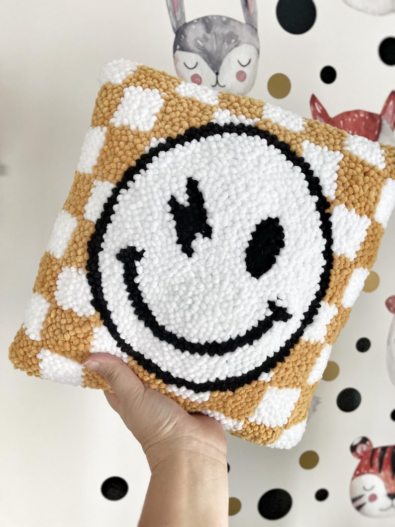 May include: A square, handmade pillow featuring a large smiley face design. The pillow has a white smiley face with black details on a checkerboard pattern of white and gold. The pillow is a decorative item.