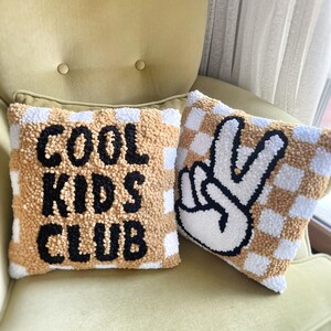 May include: Two decorative throw pillows with a checkerboard pattern in tan and white. One pillow has the words "COOL KIDS CLUB" in black. The other pillow features a black and white peace sign.