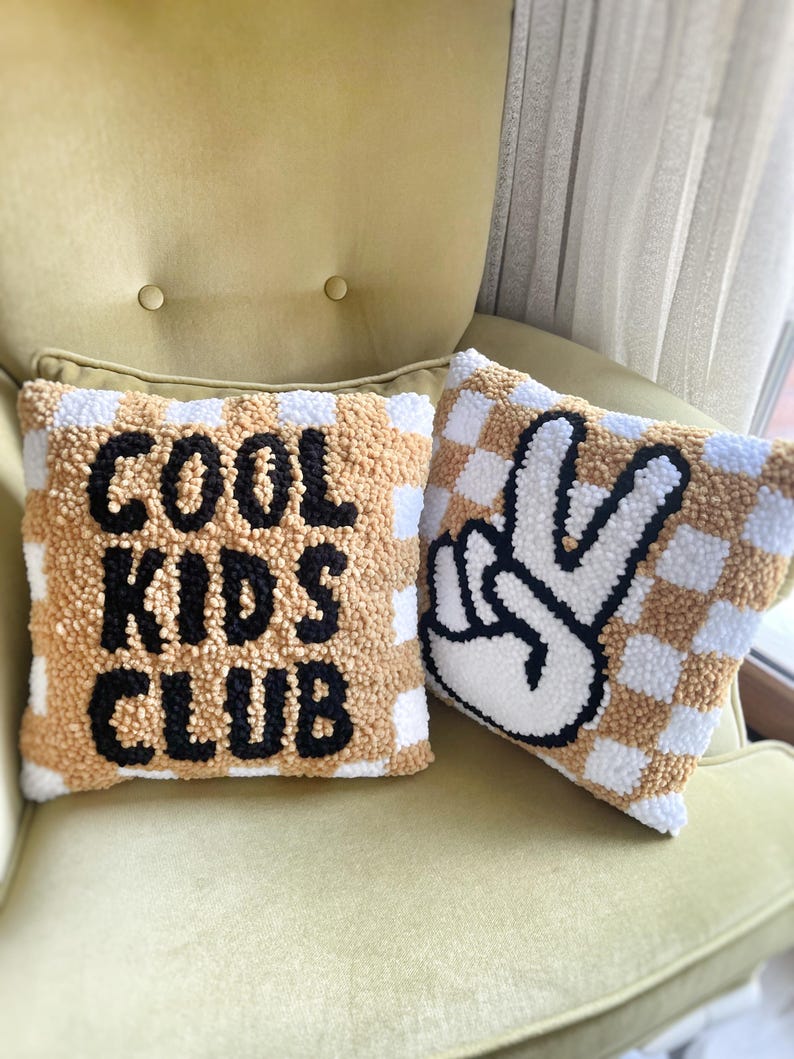 May include: Two decorative throw pillows with a checkerboard pattern in tan and white. One pillow has the words "COOL KIDS CLUB" in black. The other pillow features a black and white peace sign.