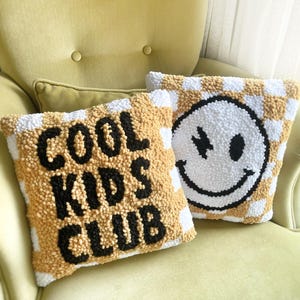May include: Two decorative throw pillows with a checkerboard pattern in white and gold. One pillow has the text "COOL KIDS CLUB" in black. The other pillow features a smiley face with a stitched design.