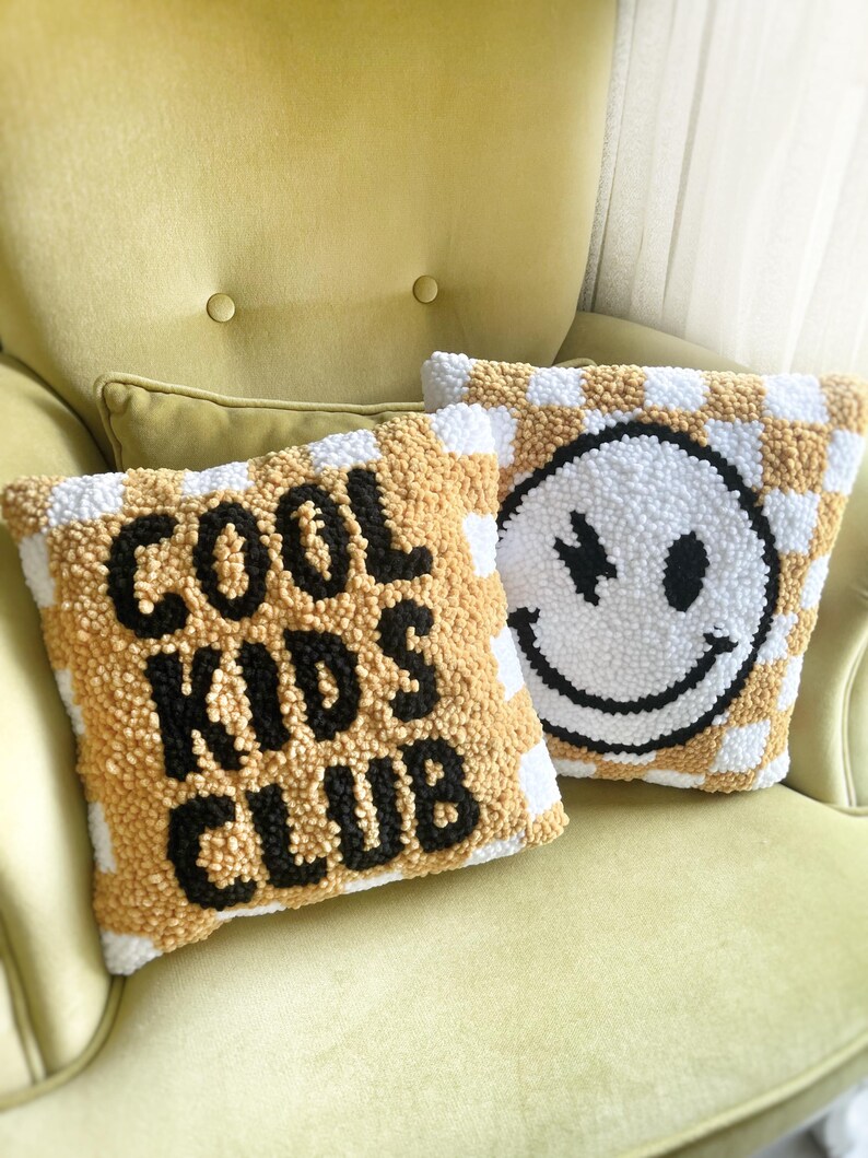 May include: Two decorative throw pillows with a checkerboard pattern in white and gold. One pillow has the text "COOL KIDS CLUB" in black. The other pillow features a smiley face with a stitched design.