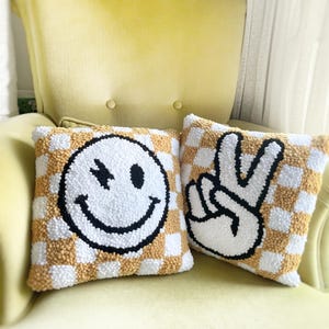 May include: Two square throw pillows with a checkerboard pattern in white and tan. One pillow features a winking smiley face, the other a peace sign, both in black and white. The pillows are on a yellow chair.