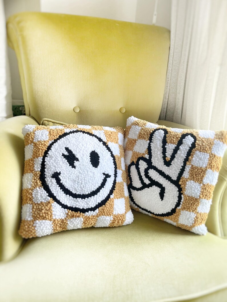 May include: Two square throw pillows with a checkerboard pattern in white and tan. One pillow features a winking smiley face, the other a peace sign, both in black and white. The pillows are on a yellow chair.