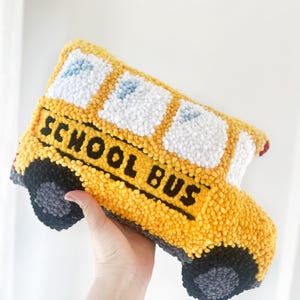 Puede incluir: Una almohada de felpa con forma de autob&uacute;s escolar amarillo con ruedas y ventanas negras. Las palabras "SCHOOL BUS" est&aacute;n bordadas en negro en el lateral del autob&uacute;s.