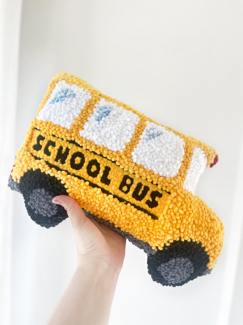 Puede incluir: Una almohada de felpa con forma de autob&uacute;s escolar amarillo con ruedas y ventanas negras. Las palabras "SCHOOL BUS" est&aacute;n bordadas en negro en el lateral del autob&uacute;s.