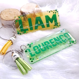 May include: Two keychains with green and yellow glitter resin tags. The tags have the names Liam and Lourenco written in green lettering. Each keychain has a silver ring and a tassel, one yellow and one green.